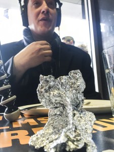 Dr Neil Verma sits at a café table wearing headphones, an empty plate which once held a slice of cake placed in front of him. In the foreground, the Frank Gehry Commemorative Wing can be seen; it suspiciously resembles a crumpled piece of aluminium foil. 